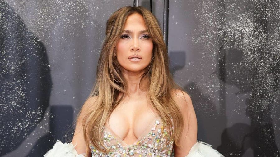A woman with long highlighted hair wearing a white ruffled sundress against a sunlit, breezy outdoor backdrop