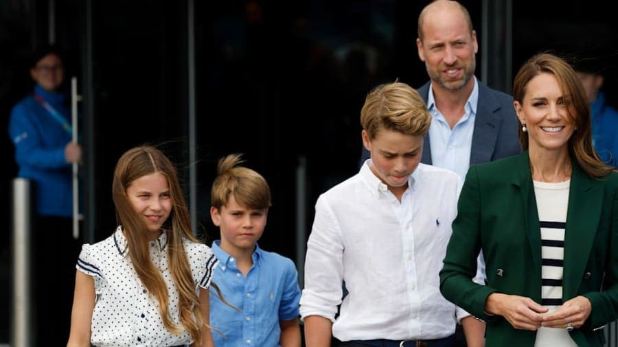 A family of five in smart-casual clothing — woman in dark green blazer, man in navy suit, two boys in button-down shirts, girl in polka-dot dress — seated in sports stadium stands