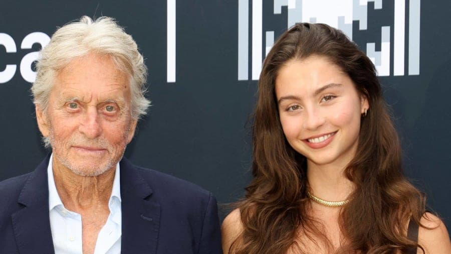 An elderly man wearing a navy suit and a young woman wearing a black strapless top and wide-leg trousers against a red carpet backdrop at an evening awards ceremony