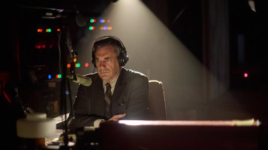 A man in 1970s clothing standing at a radio broadcasting desk against a dimly lit studio backdrop