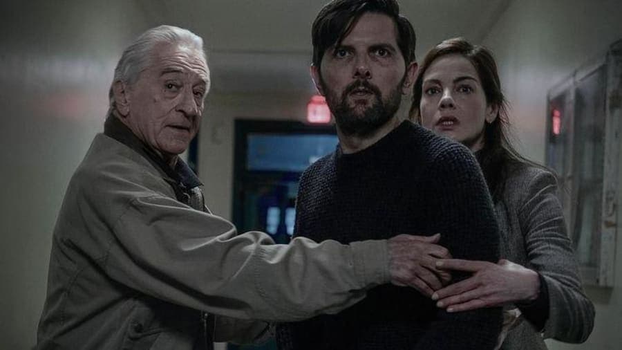Three actors in a tense scene — a grey-haired older man on the left, a dark-haired man in the centre, and a woman on the right — appearing to confer urgently in what looks like an indoor setting