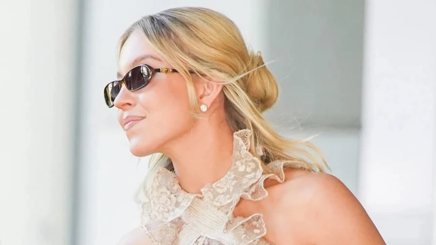 A young blonde woman wearing a sheer floral lace cropped top layered over a peach tank top, paired with sand-coloured wide-leg shorts and nude heeled pumps, photographed outdoors in a European city setting while carrying a small gold handbag