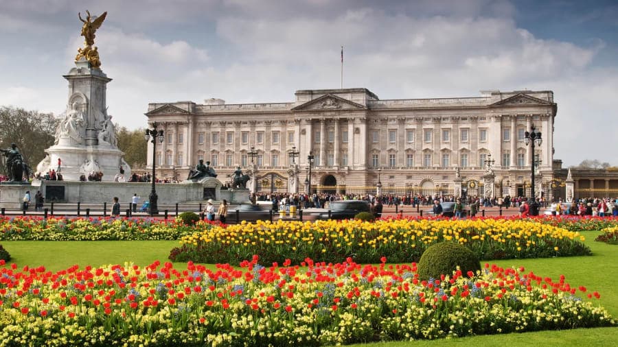 The ornate stone facade and gilded gates of a grand royal palace in London, with uniformed guards visible at the entrance