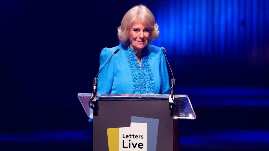 A woman in a blue evening gown stands at a lectern on a stage at a large London concert hall, addressing a packed audience