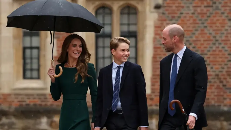 A teenage boy in a dark blazer, shirt, tie and chinos walks across a cobblestone courtyard with two adults beside him in the rain, approaching a historic stone building