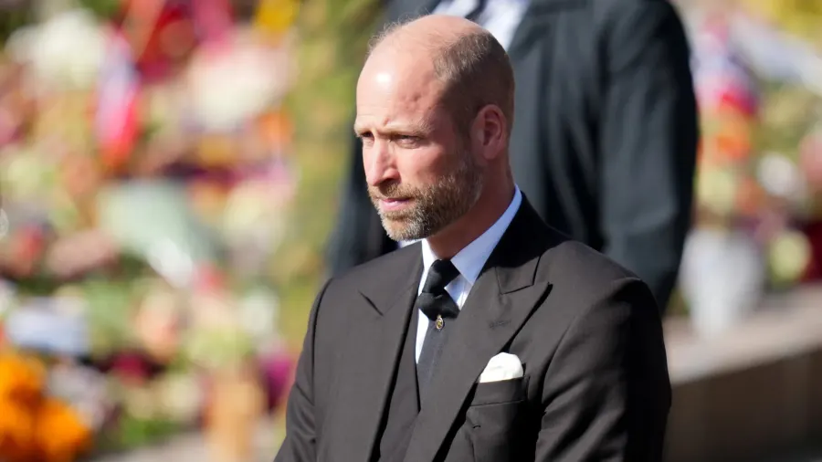 A man in a dark suit walks solemnly alongside a woman in dark formal attire during an outdoor funeral procession, with other formally dressed mourners visible behind them