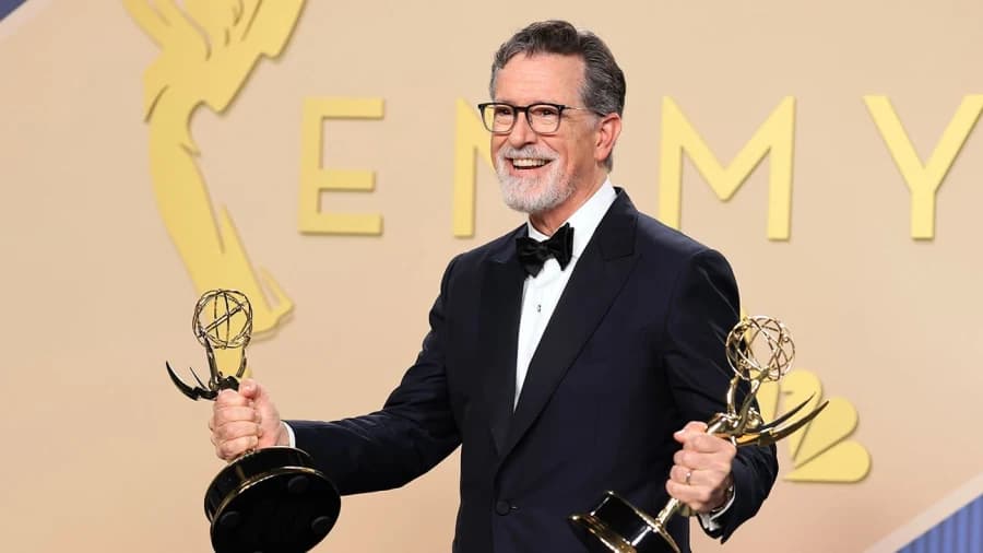A bearded man in formal evening wear stands at an awards ceremony podium, holding an Emmy statuette and speaking into a microphone on a brightly lit stage