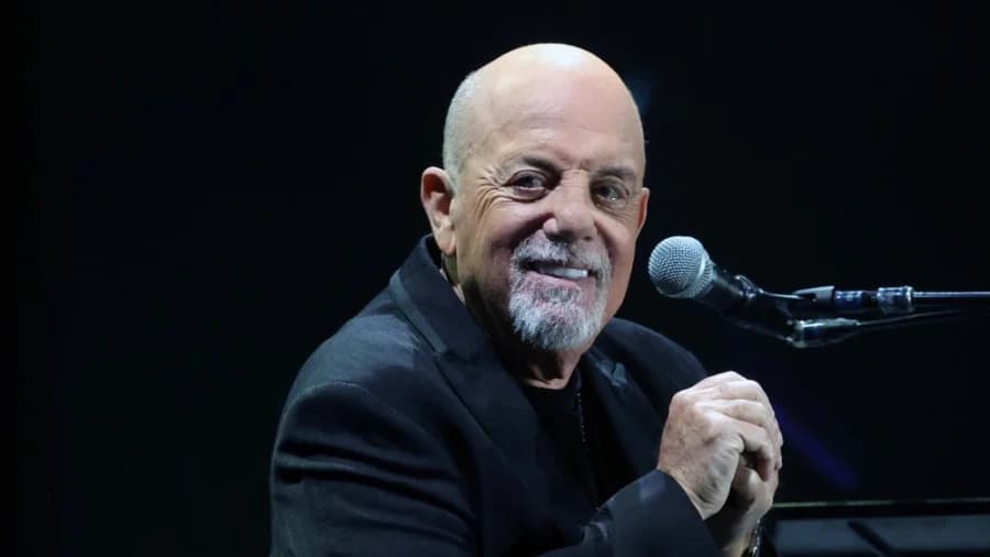 An older man with grey hair in a dark jacket speaking into a microphone on stage, hands raised, performing in front of a large audience