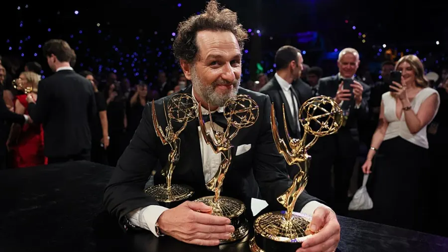 A man in formal evening wear holding two Emmy statuettes on a stage at an awards ceremony, smiling at the crowd
