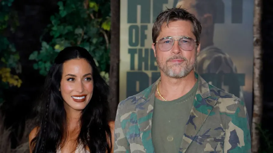 A man in a camouflage suit and aviator sunglasses and a woman in a pale pink tiered dress with lace trim walk hand-in-hand on a red carpet at an outdoor premiere venue