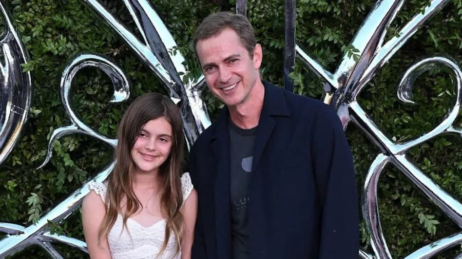 A man in a navy blazer and printed T-shirt poses on a red carpet alongside a young girl in a white lace dress, both smiling at the camera