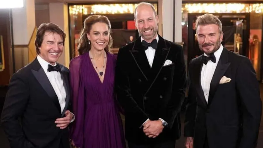 A man in a black tuxedo stands with two younger men also in black tie, alongside a woman in a floor-length plum gown and a man in a velvet evening jacket, posing together at a film premiere red carpet event inside a grand cinema foyer