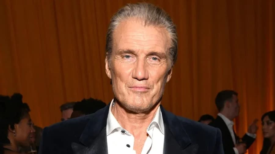 A broad-shouldered man with cropped grey hair, wearing a dark jacket, photographed at a media event against a branded backdrop