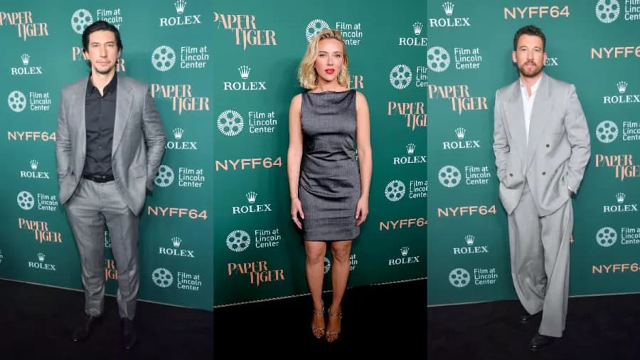 A woman in a sleeveless charcoal-gray minidress with metallic silver heels stands on a red carpet at a film festival premiere, flanked by two men in gray suits