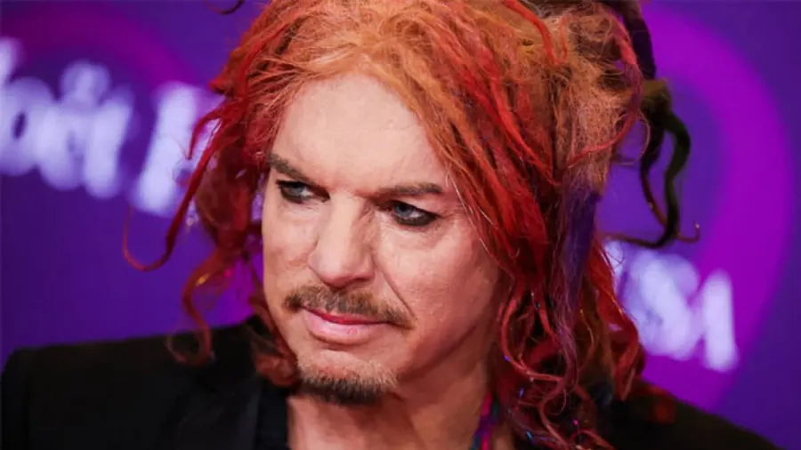 A man with curly red hair standing on a stage in front of a colourful backdrop, wearing a dark outfit