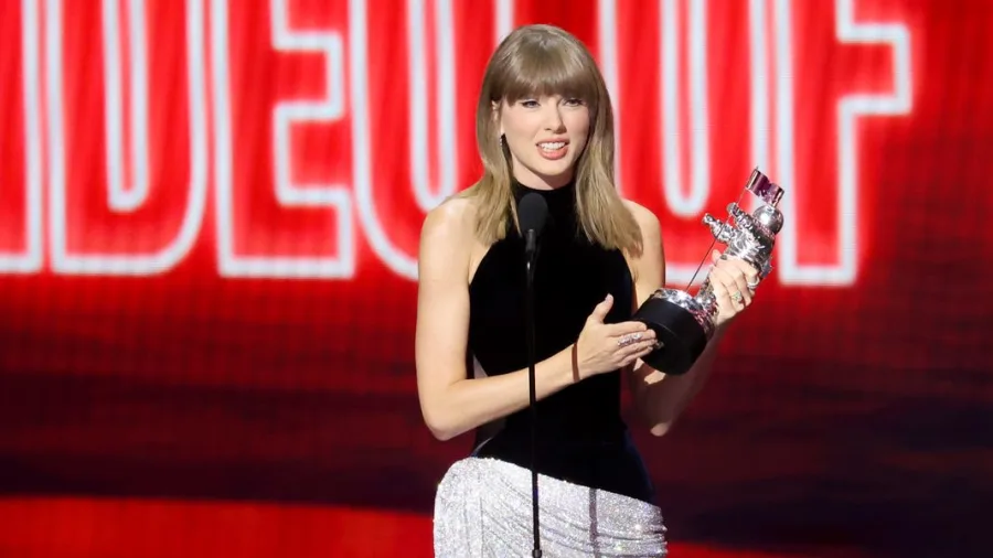 A woman in an elaborate stage outfit stands at a podium on a large awards show stage, holding a trophy and speaking into a microphone, surrounded by dramatic lighting