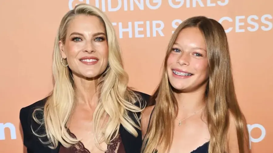 A blonde woman in a navy pantsuit stands alongside a young blonde girl in a navy mini dress on a red carpet, both smiling at the camera