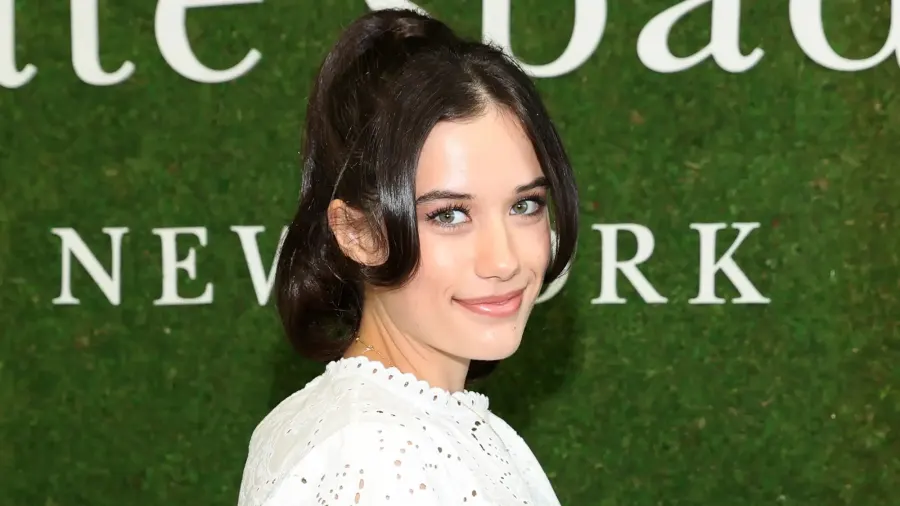 A young woman with dark hair walks a fashion show runway wearing an oversized dark green blazer over a patterned blouse with dark trousers, amid bright runway lighting